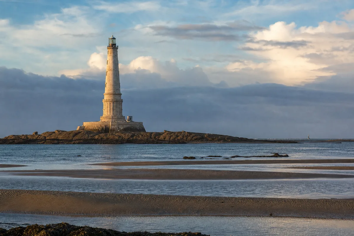 Phare ancien en pierre dressé sur un plateau rocheux à l&rsquo;embouchure d&rsquo;un estuaire, mer calme au premier plan